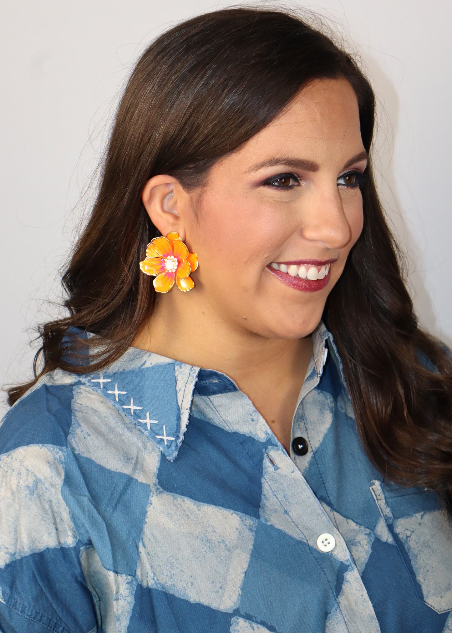 Woman wearing a blue and white checkered shirt with neon orange floral earrings against a plain background