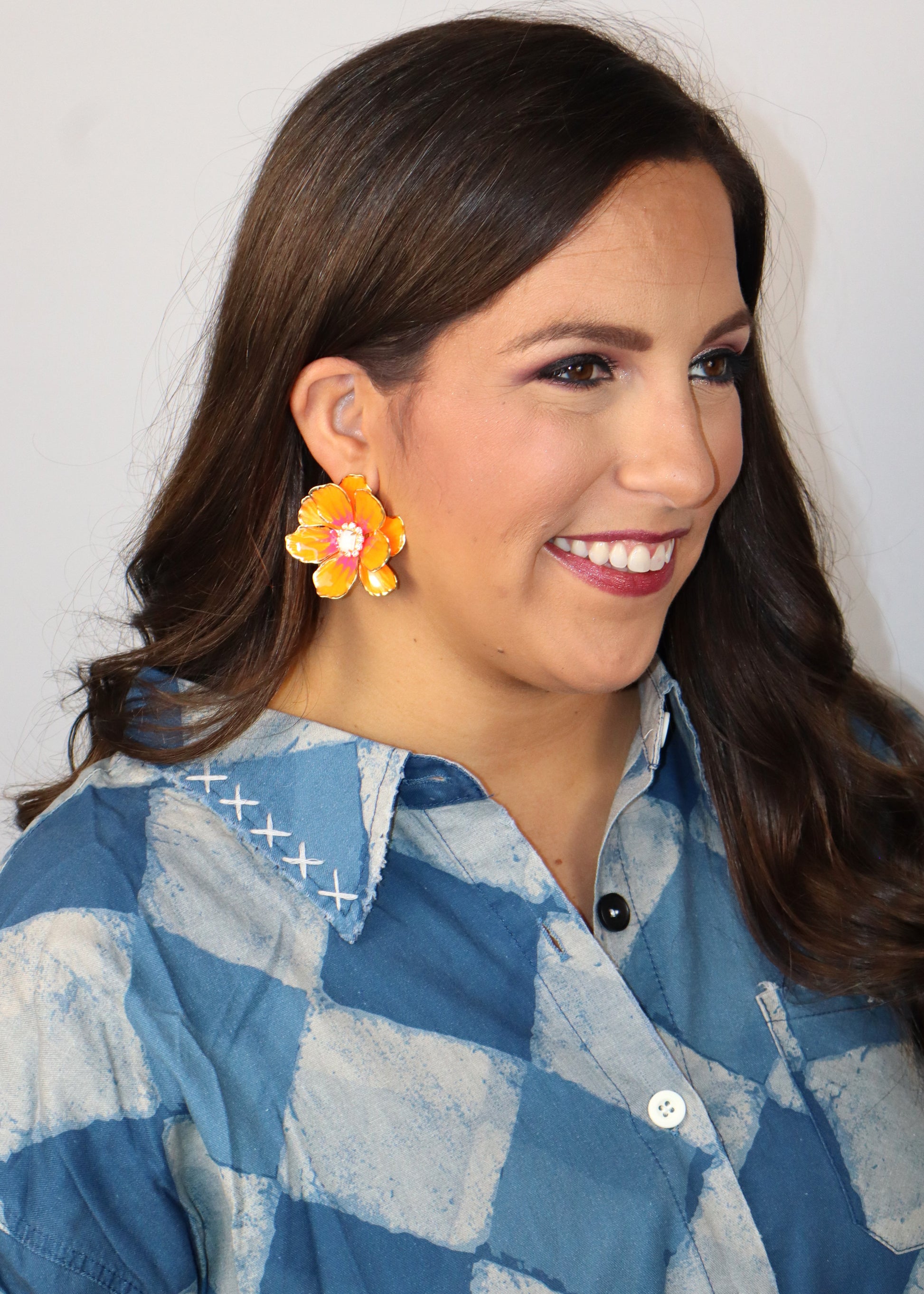 Woman wearing a blue and white checkered shirt with neon orange floral earrings against a plain background