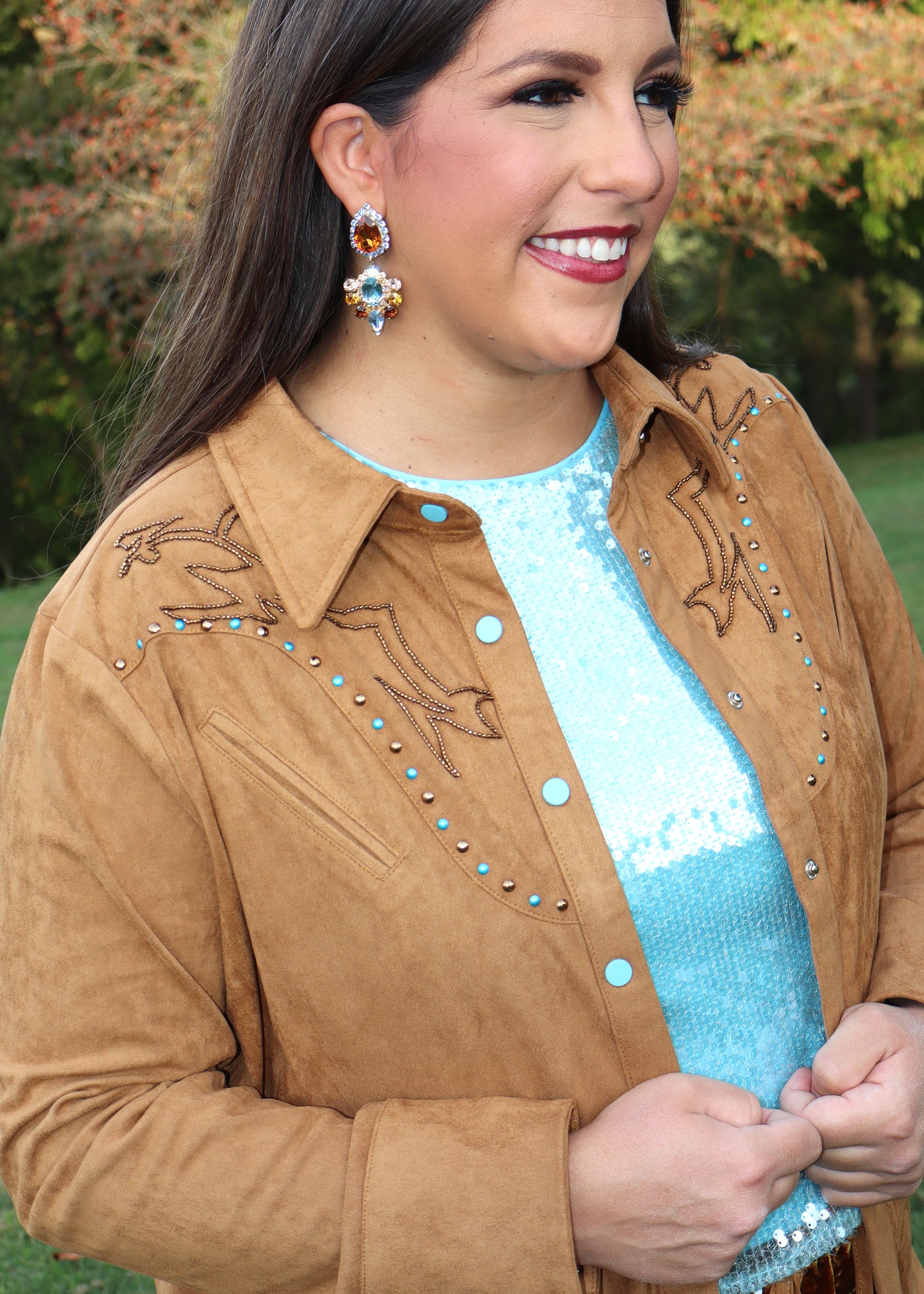 Woman wearing a camel-colored western-style, suede shirt and blue sequin shirt standing outside near trees