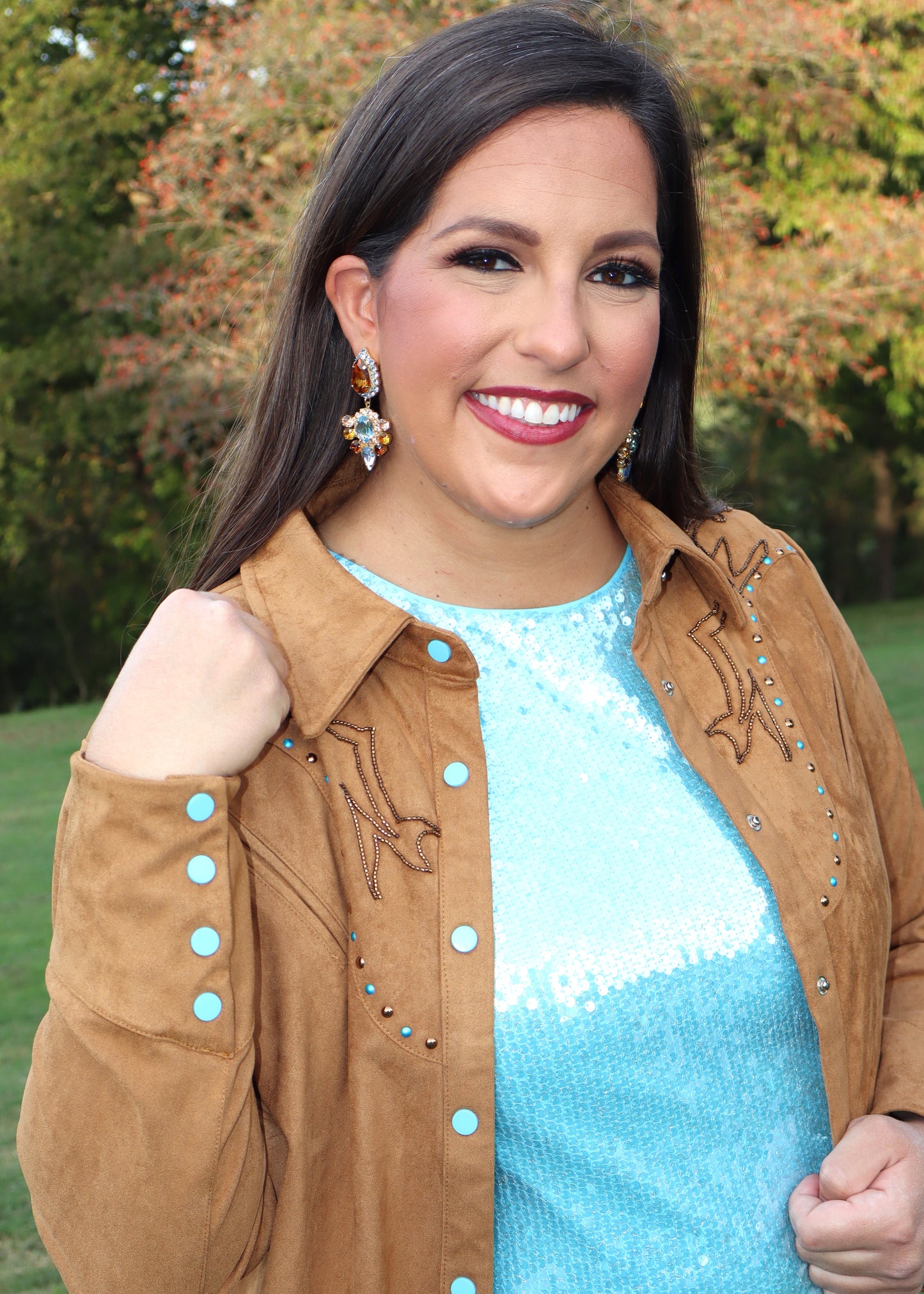 Woman wearing a camel-colored western-style, suede shirt and blue sequin shirt standing outside near trees