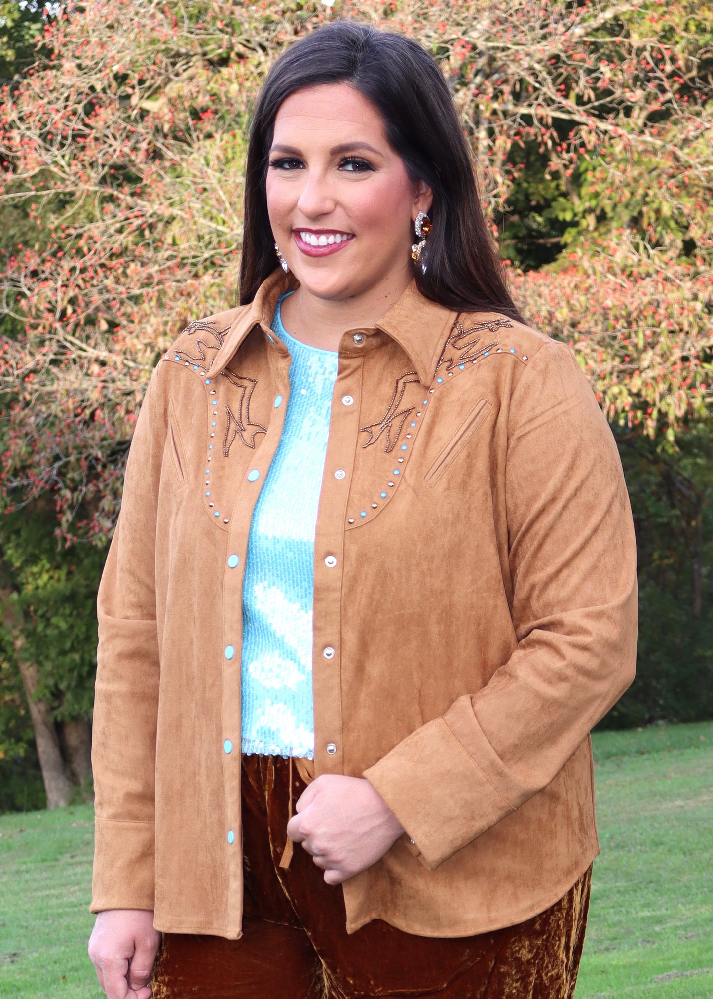Woman wearing a camel-colored western-style, suede shirt and blue sequin shirt standing outside near trees
