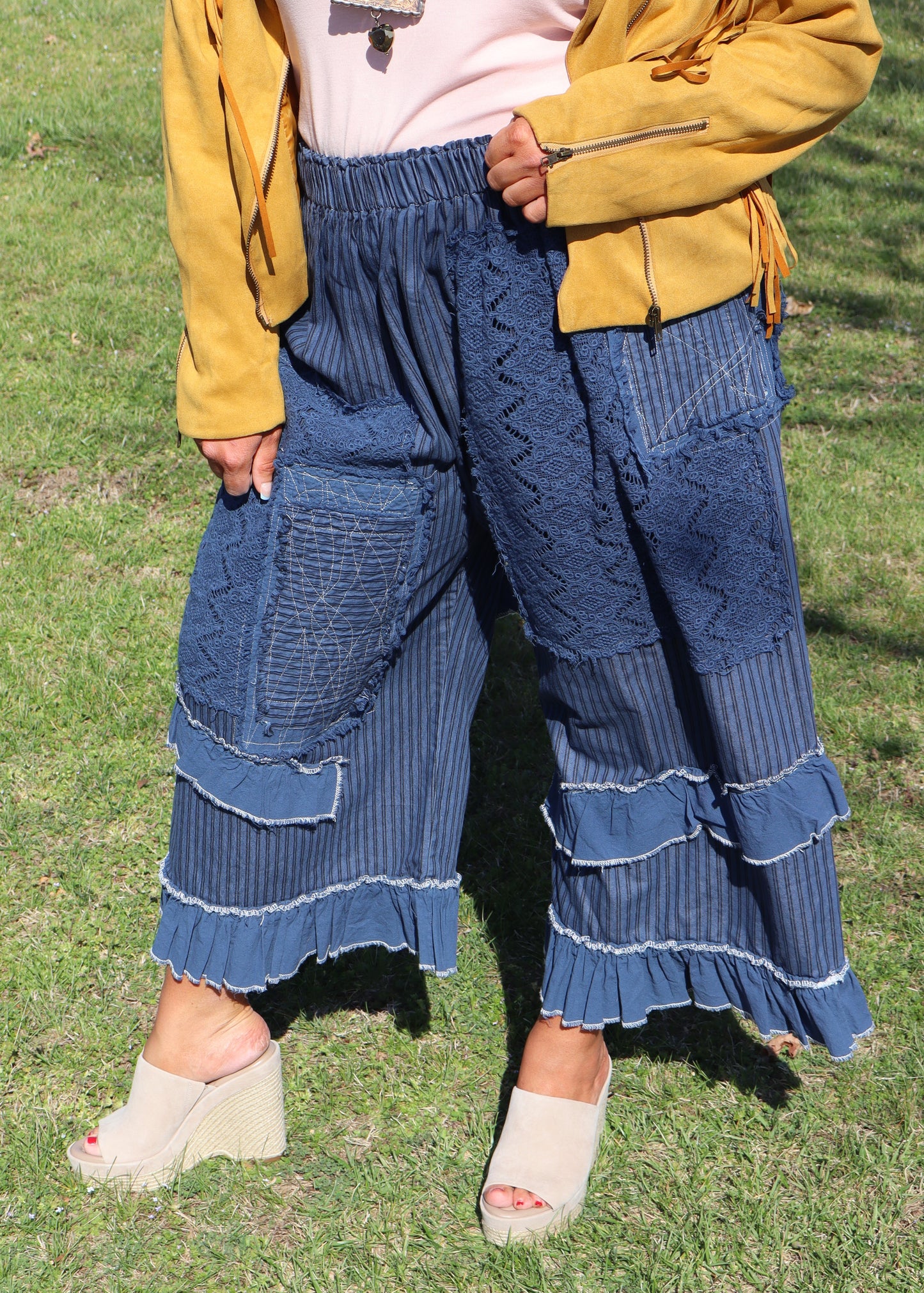 Woman in a mustard yellow suede jacket, pink top, and blue ruffle pants standing in a grassy field.