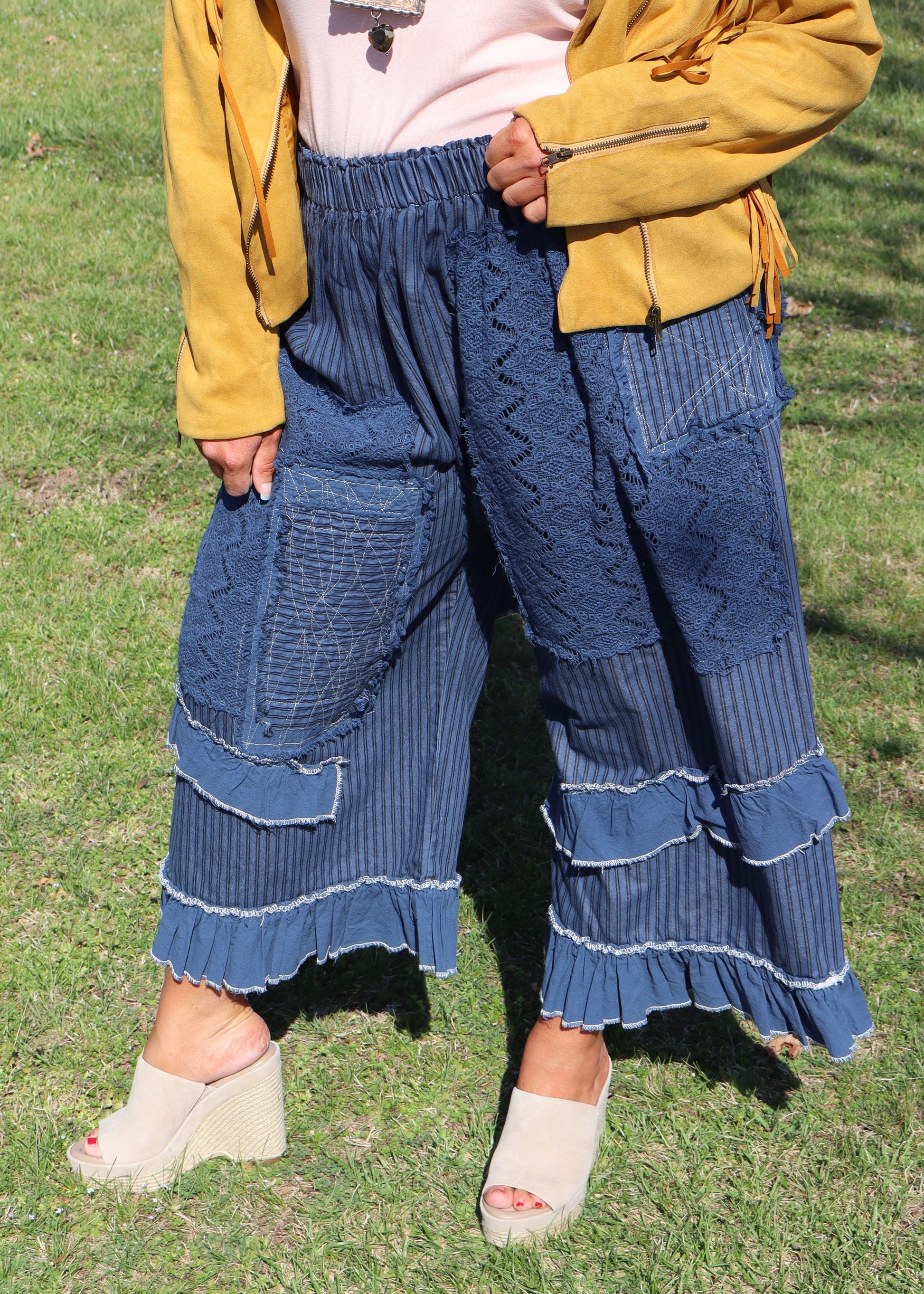 Woman in a mustard yellow suede jacket, pink top, and blue ruffle pants standing in a grassy field.