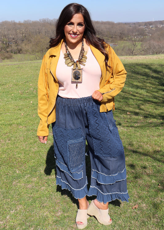 Woman in a mustard yellow suede jacket, pink top, and blue ruffle pants standing in a grassy field.