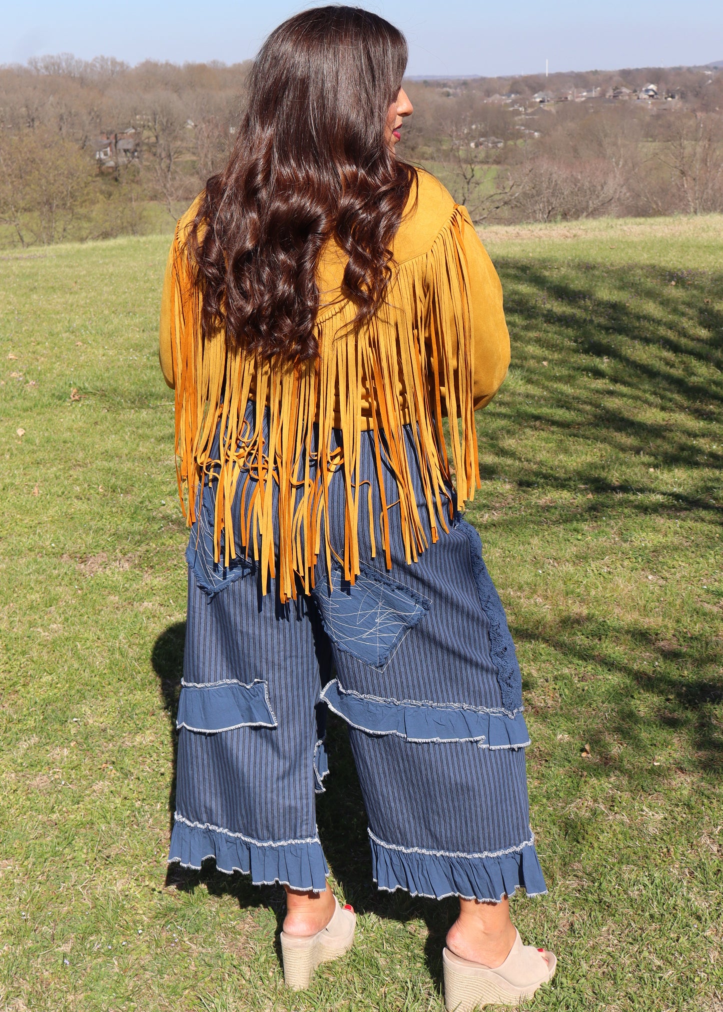 Woman in a mustard yellow suede fringe jacket and blue ruffle pants standing in a grassy field.