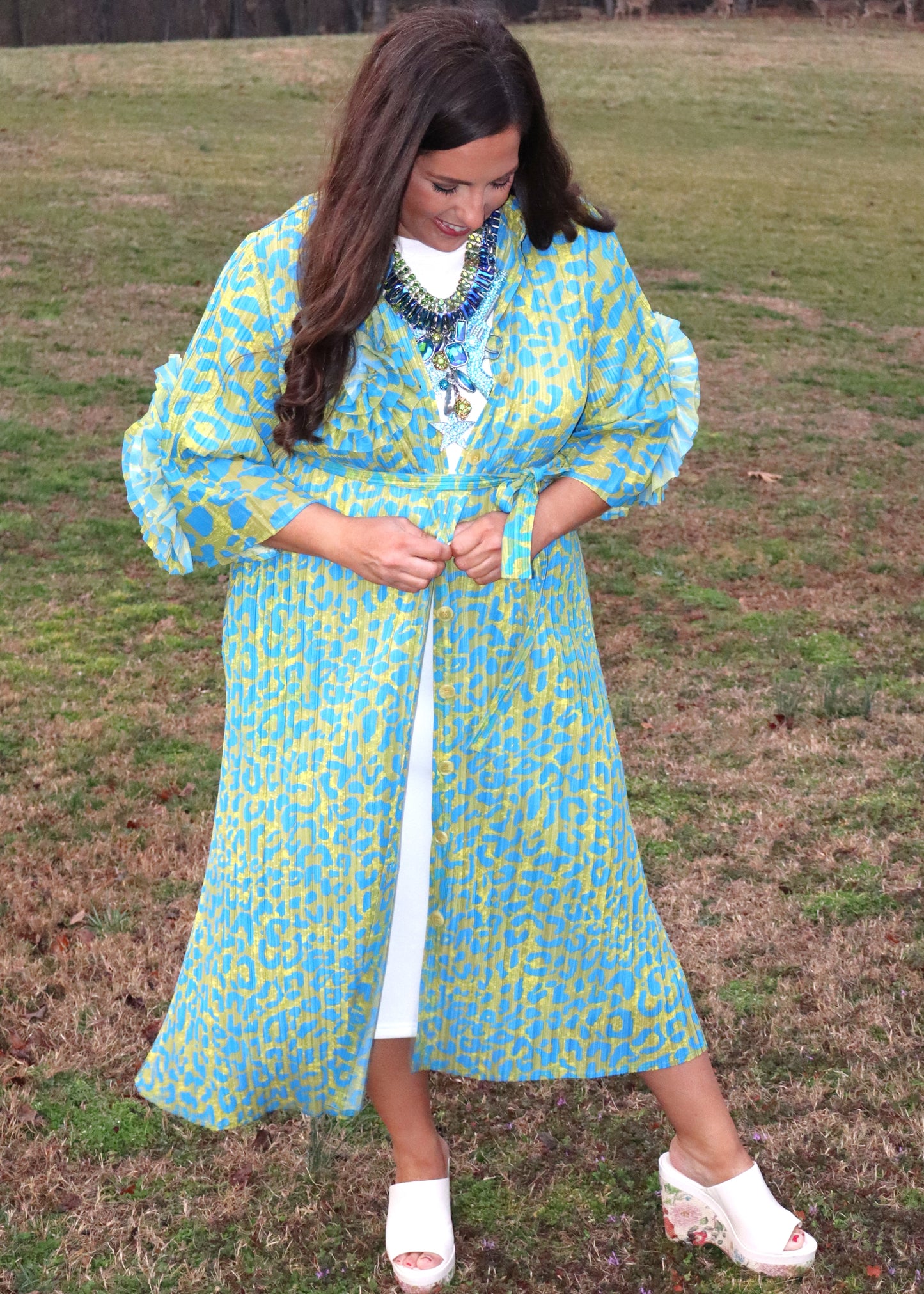 Woman wearing a leopard print, green and blue button-up kimono dress with large 3D floral details on the sleeves and a waist tie, standing outside. 