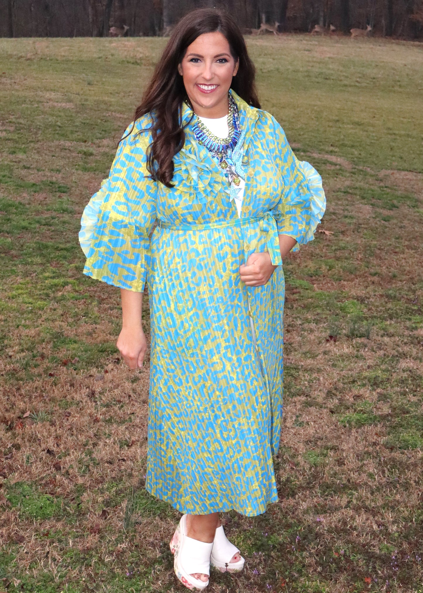 Woman wearing a leopard print, green and blue button-up kimono dress with large 3D floral details on the sleeves and a waist tie, standing outside. 