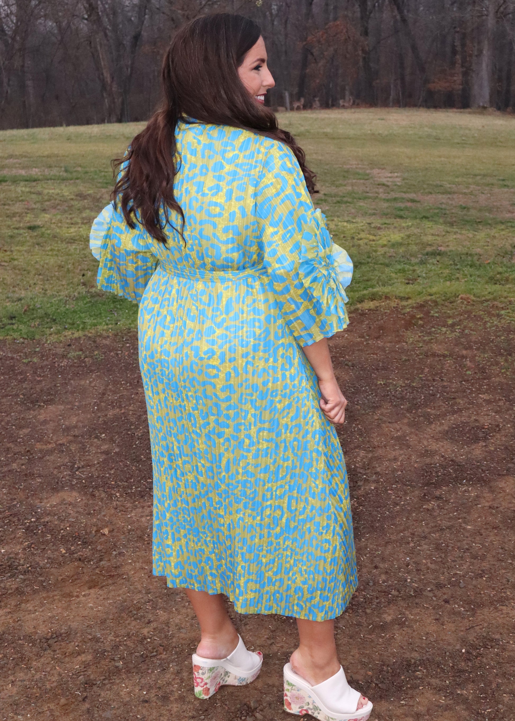 Woman wearing a leopard print, green and blue button-up kimono dress with large 3D floral details on the sleeves and a waist tie, standing outside. 