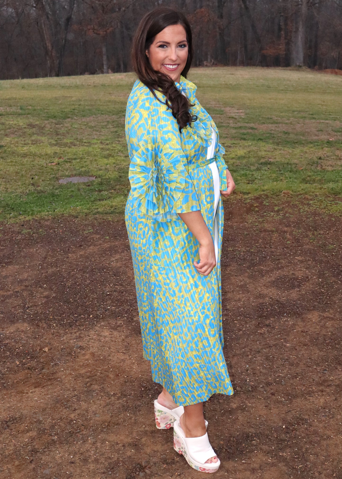 Woman wearing a leopard print, green and blue button-up kimono dress with large 3D floral details on the sleeves and a waist tie, standing outside. 