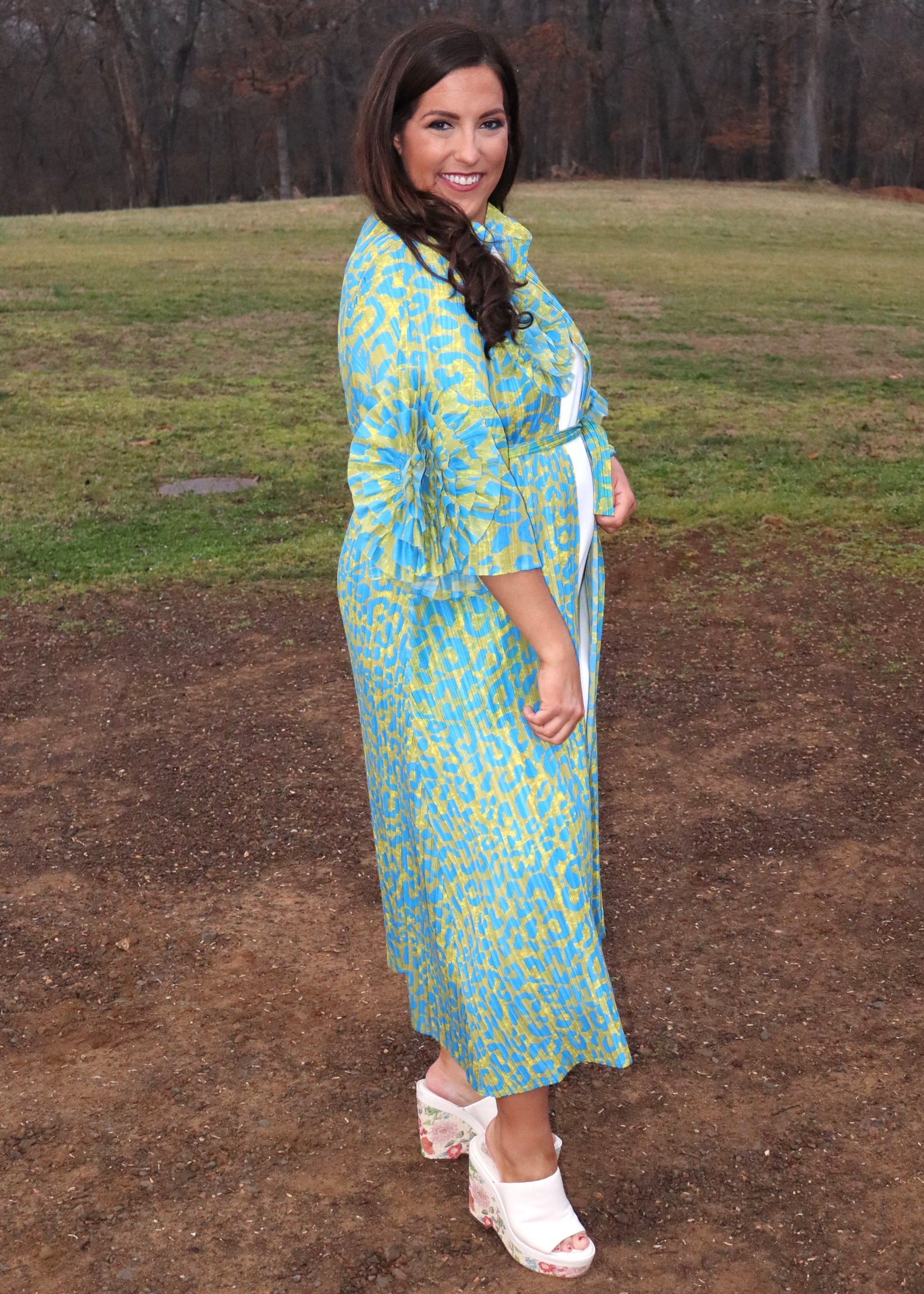 Woman wearing a leopard print, green and blue button-up kimono dress with large 3D floral details on the sleeves and a waist tie, standing outside. 