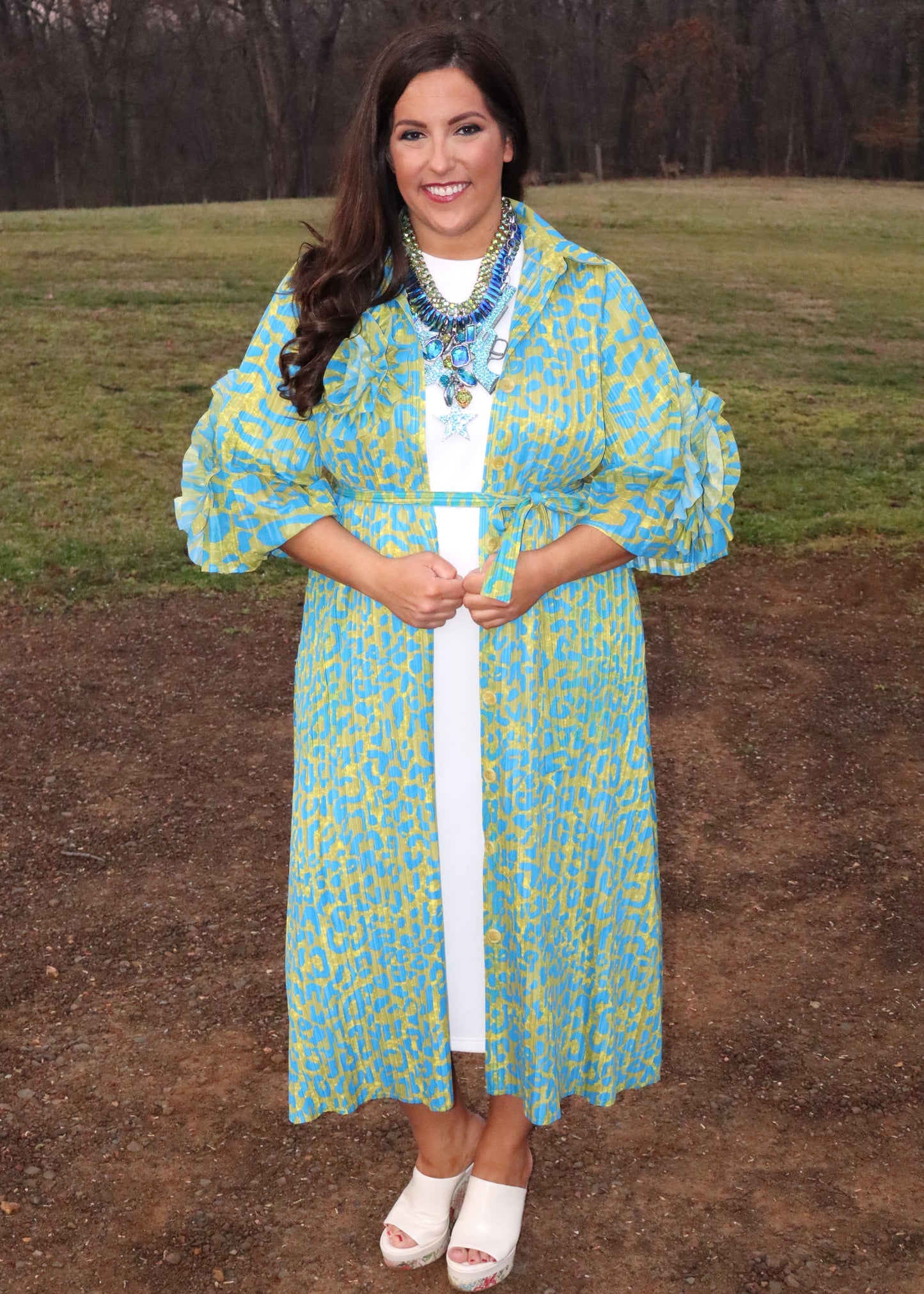 Woman wearing a leopard print, green and blue button-up kimono dress with large 3D floral details on the sleeves and a waist tie, standing outside. 