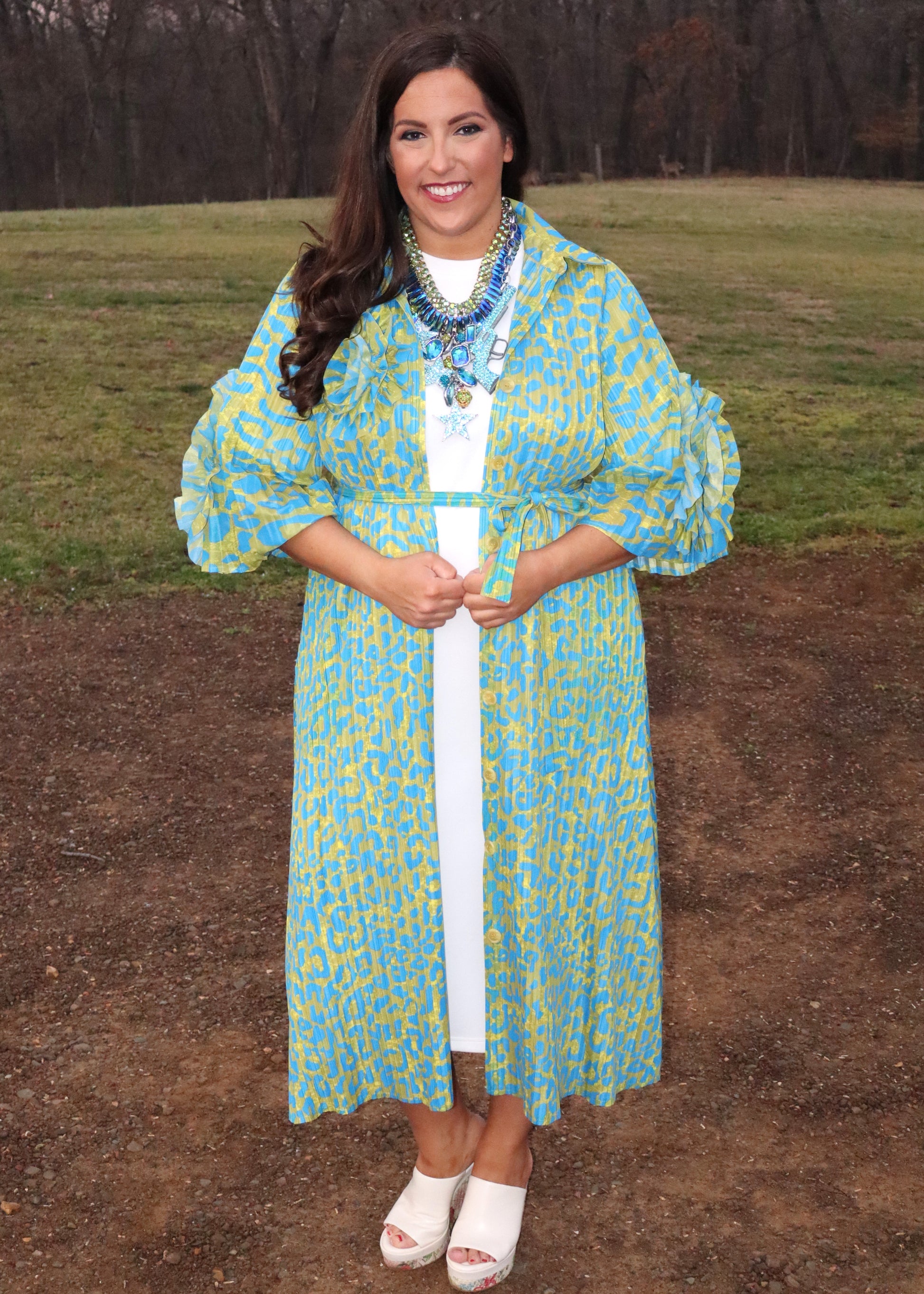 Woman wearing a leopard print, green and blue button-up kimono dress with large 3D floral details on the sleeves and a waist tie, standing outside. 