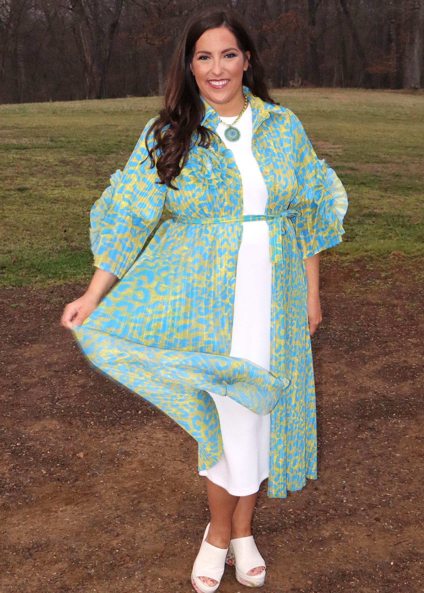 Woman wearing a leopard print, green and blue button-up kimono dress with large 3D floral details on the sleeves and a waist tie, standing outside. 