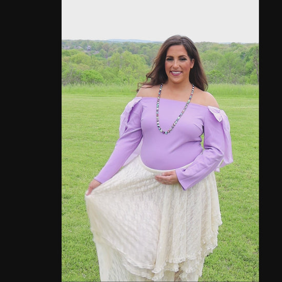 Video of woman in an off-the-shoulder light purple top and cream lace skirt standing in a grassy field.