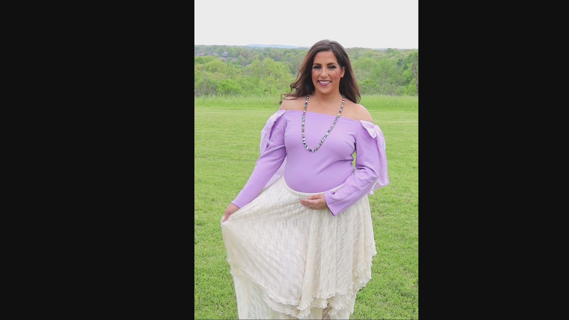 Video of woman in an off-the-shoulder light purple top and cream lace skirt standing in a grassy field.