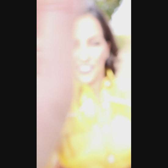 Video of woman wearing a yellow, shimmery blouse and bronze pleated skirt outdoors with trees in the background