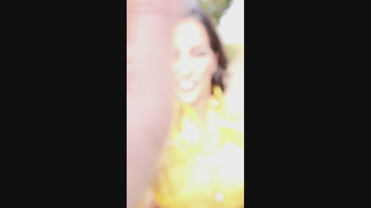 Video of woman wearing a yellow, shimmery blouse and bronze pleated skirt outdoors with trees in the background