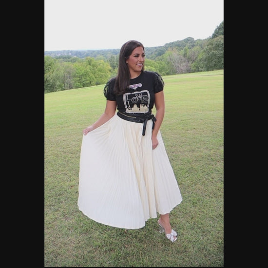 Video of woman wearing a black t-shirt with perfume bottle design and an ivory, pleated skirt in an outdoor setting showing how the skirt flows.