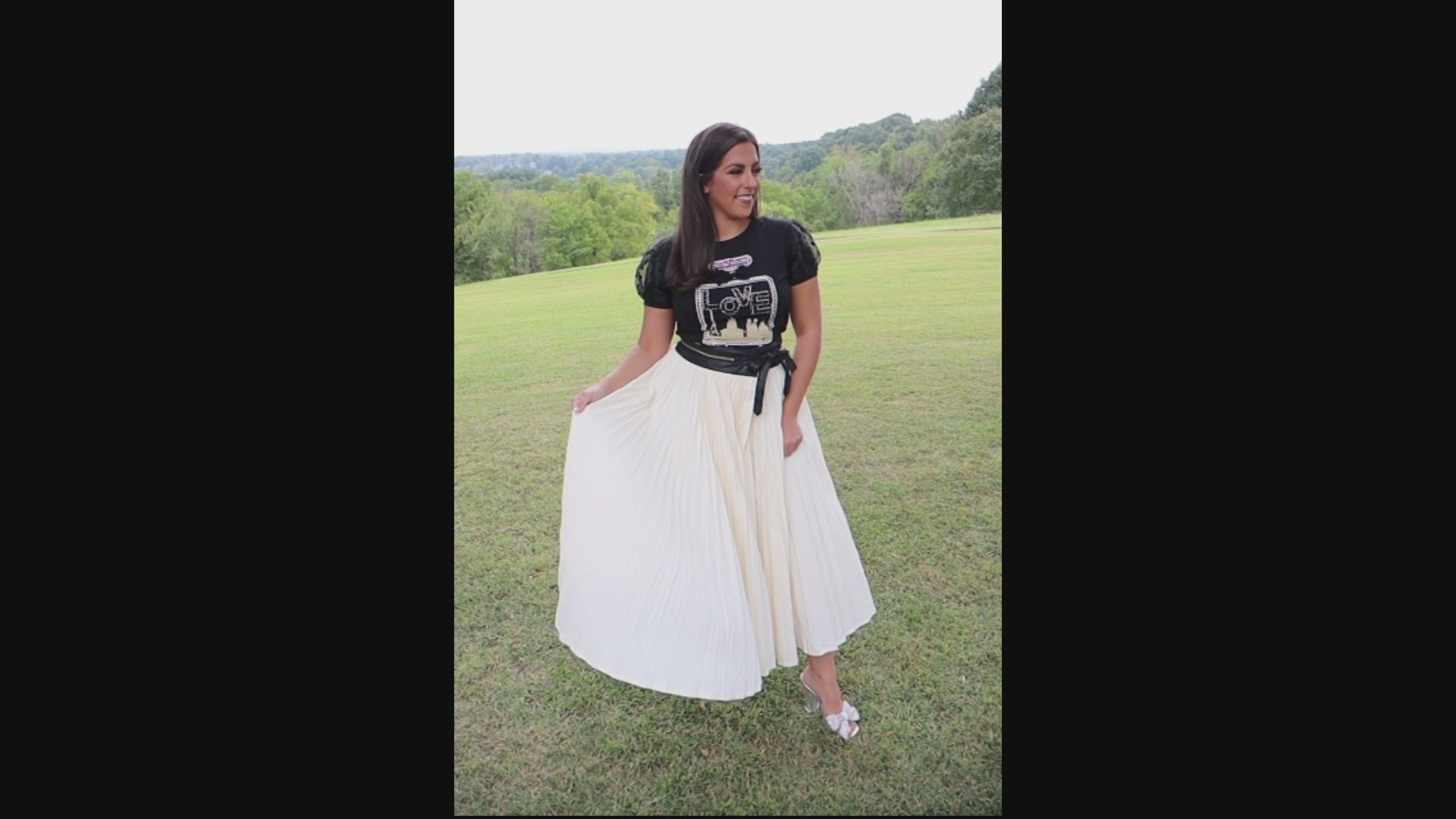 Video of woman wearing a black t-shirt with perfume bottle design and an ivory, pleated skirt in an outdoor setting showing how the skirt flows.